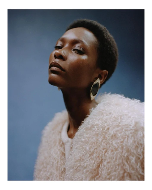 Portrait of an African-American woman in a peach faux-fur coat against a blue background. She wears geometric earrings and looks upward.
