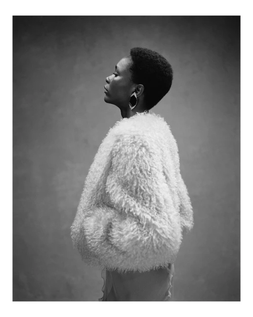 Monochrome portrait of a Black woman in profile, wearing statement earrings and a shaggy white coat against a textured grey backdrop.