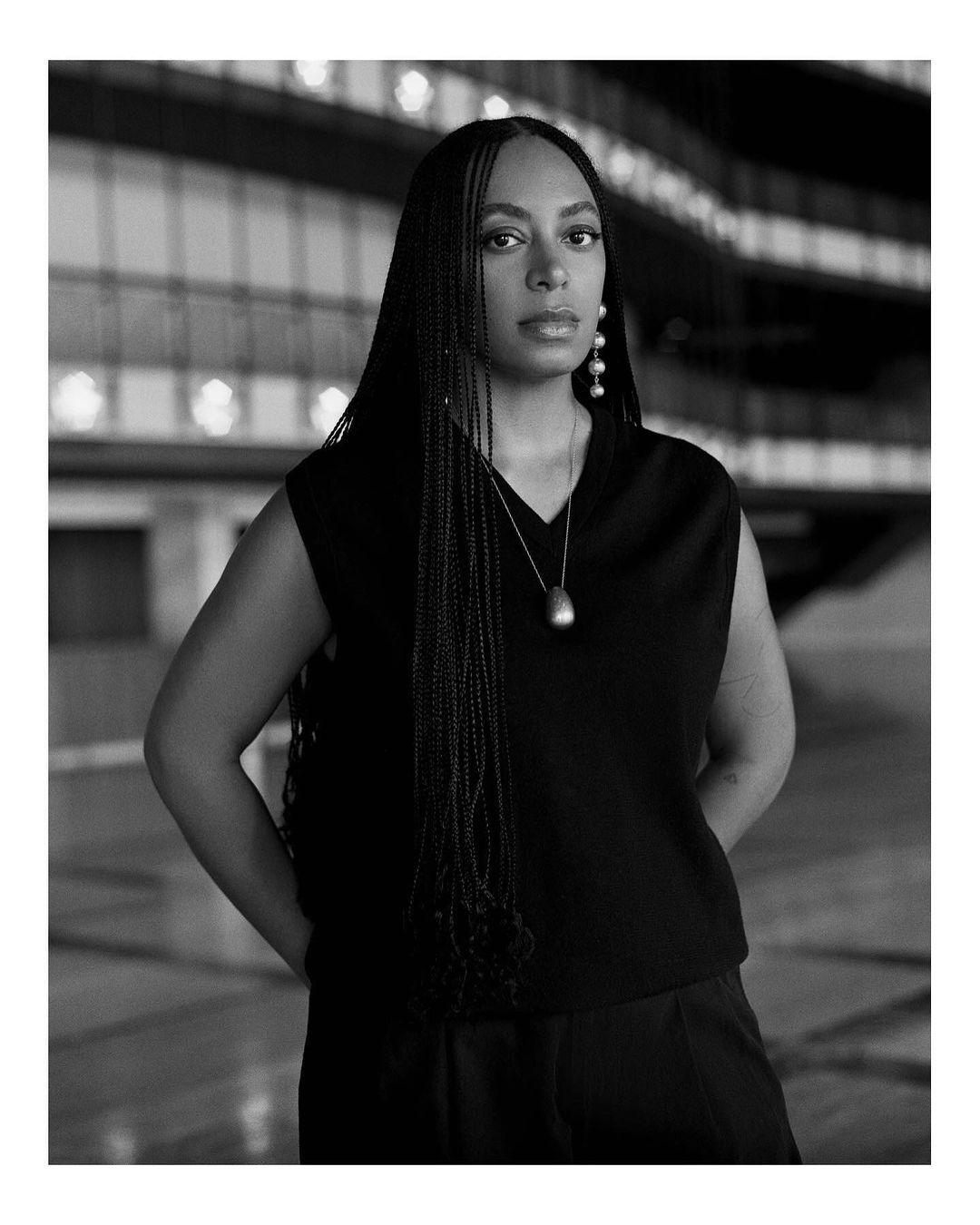 Black and white portrait of a woman with long braided hair wearing a dark top and trousers, posing in an indoor location.