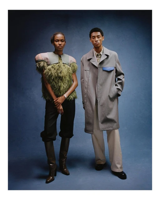 Two fashion models pose in studio against a blue backdrop. Woman wears an olive green feathered top and black trousers with boots. Man wears a trench coat.