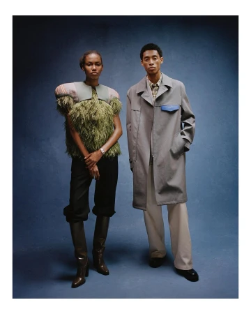 Two fashion models pose in studio against a blue backdrop. Woman wears an olive green feathered top and black trousers with boots. Man wears a trench coat.