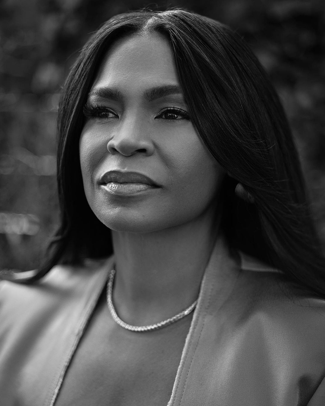 Black and white portrait of a woman with long dark hair wearing a blazer and necklace, looking off to the side with a serene expression.