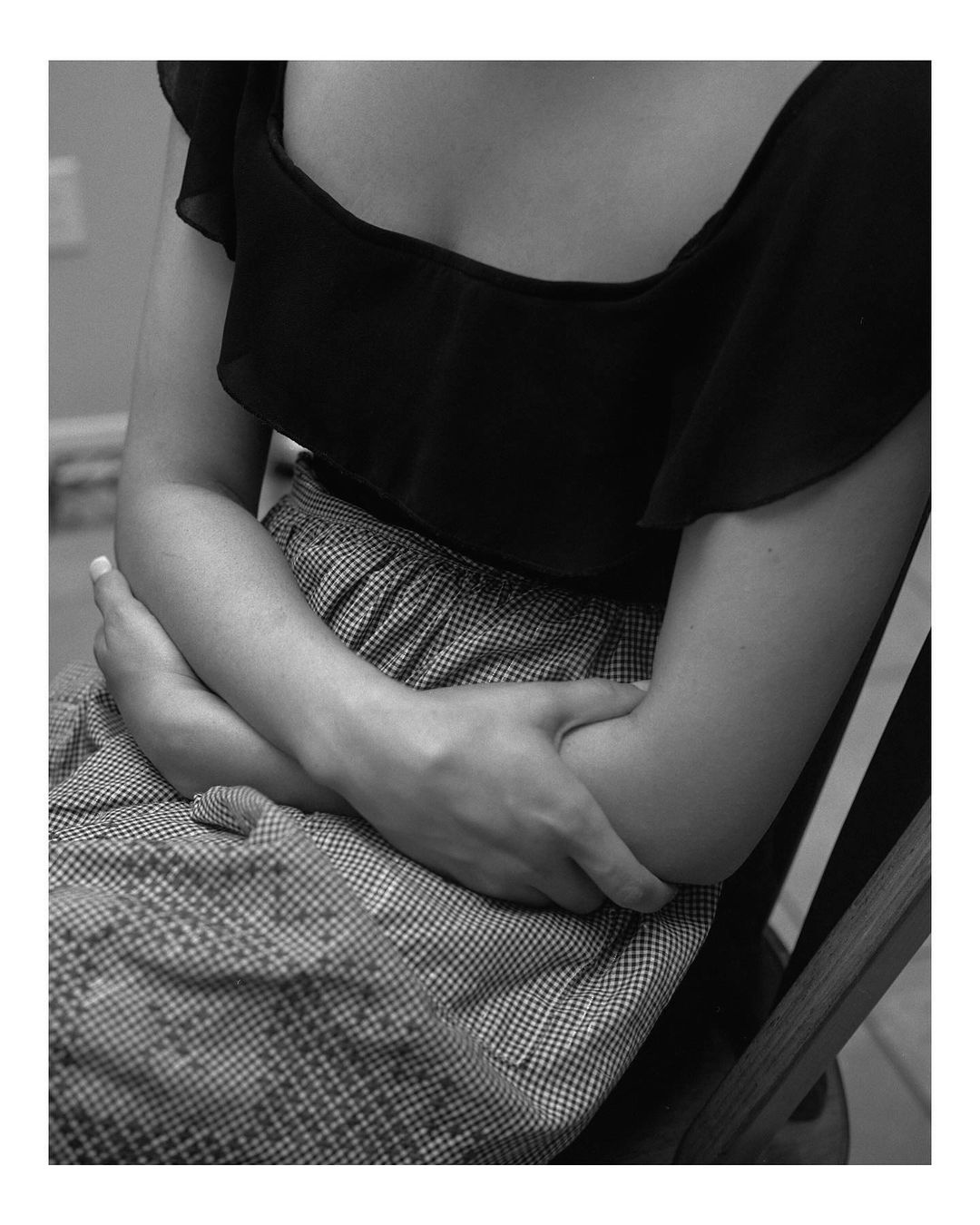 Black and white portrait of a woman with folded arms wearing a dark, ruffled top and patterned skirt while sitting on a wooden chair.