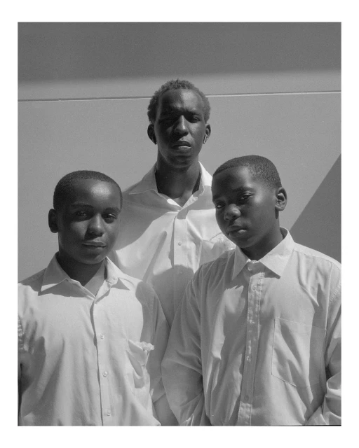 Black-and-white portrait of three African American teenage boys in white shirts, standing in front of a light-colored wall.
