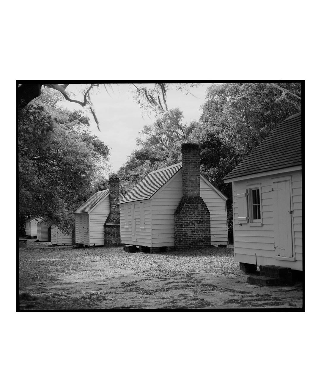 Monochrome photograph of a row of small, white, wooden buildings with brick chimneys, set against a backdrop of leafy trees.