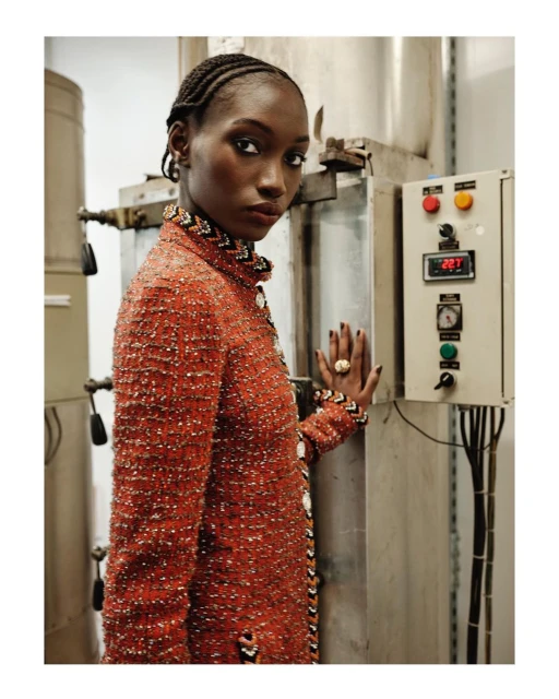 A Black woman in a patterned orange jacket stands next to industrial equipment in a fashion editorial shot with neutral lighting indoors.