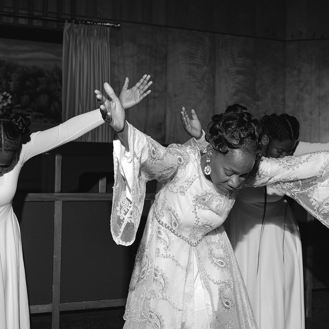 Three African-American women in formal gowns stand in a line with arms outstretched in a softly lit indoor setting.