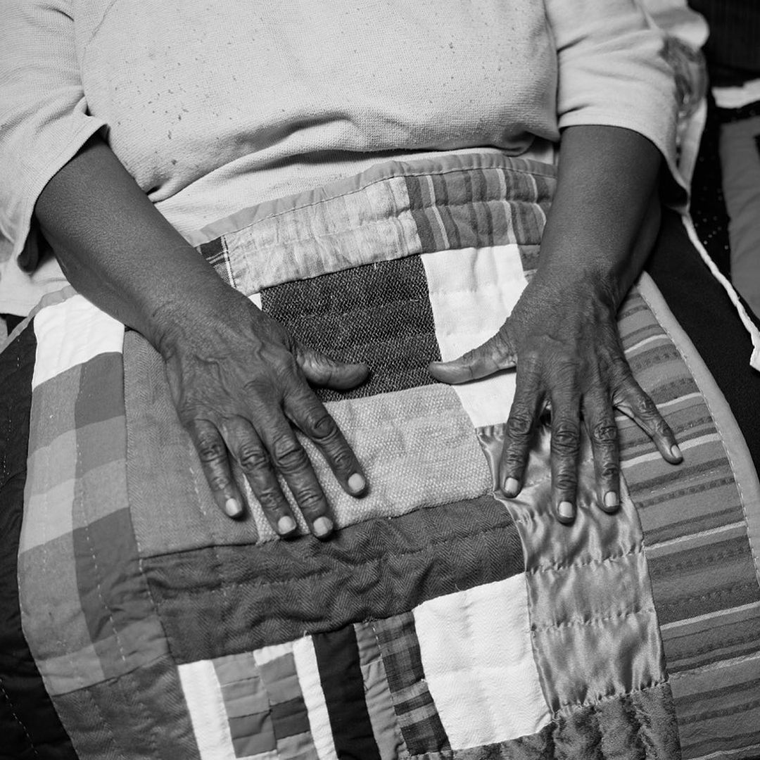 Black and white photograph features an elderly person's hands resting on a multicolored patchwork quilt covering their lap, indoors.
