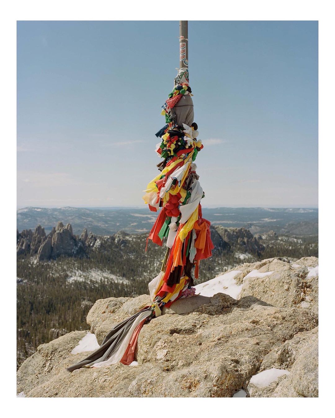 Colorful prayer flags adorn a metal pole atop a rocky, snow-dusted peak, with a panoramic mountain landscape under a clear blue sky.