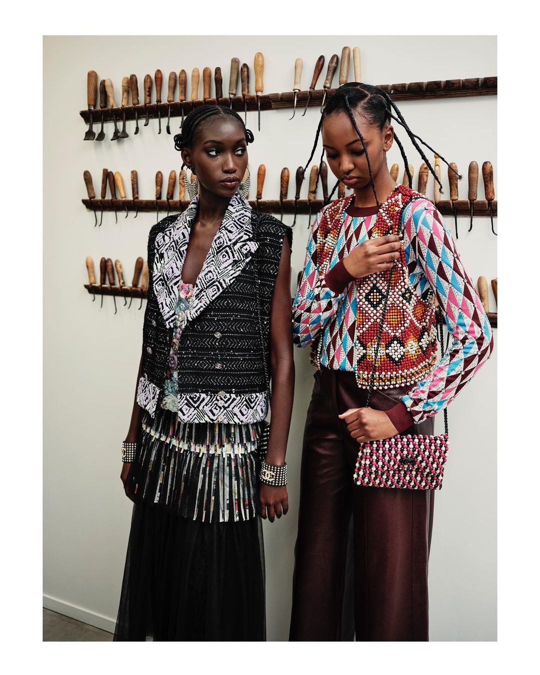 Two African American women pose indoors; one wears a black and white vest, and the other a patterned top with red pants and purse.