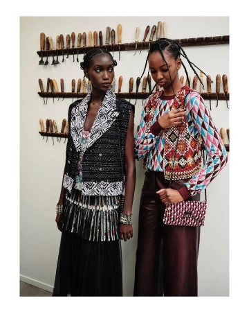 Two African American women pose indoors; one wears a black and white vest, and the other a patterned top with red pants and purse.