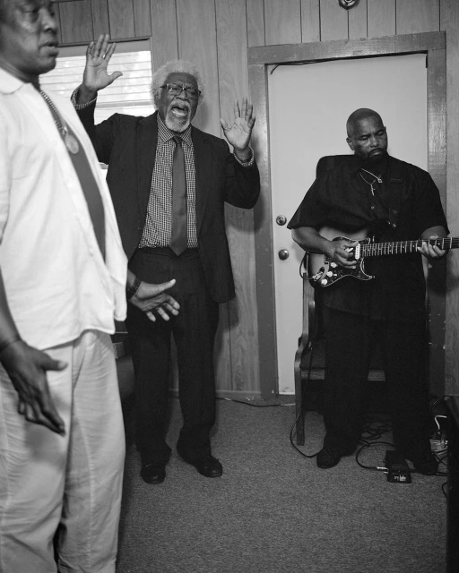 A grayscale photo shows three African-American men, one singing with raised hands, another playing a guitar, and the third listening.