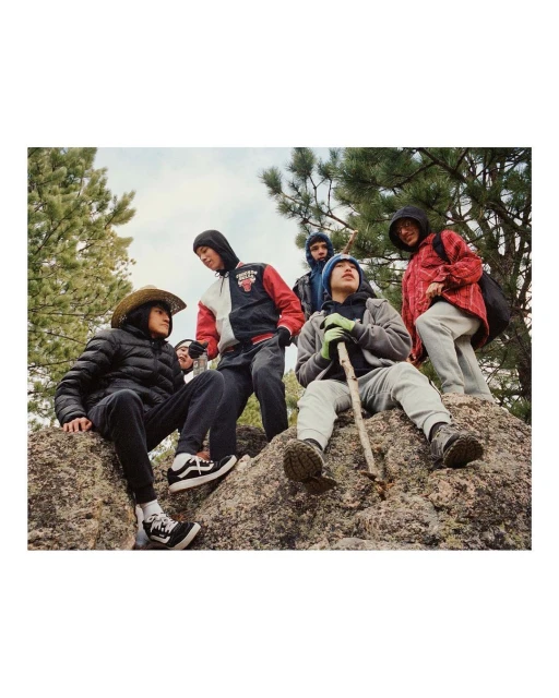Group of young men gathers on a rock outcrop in a natural setting, wearing a variety of casual jackets, hoodies, jeans, and sneakers.