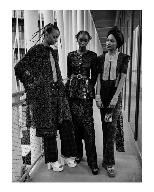 Three fashion models in a black and white photo, showcasing patterned outfits with platform shoes in an indoor corridor setting.