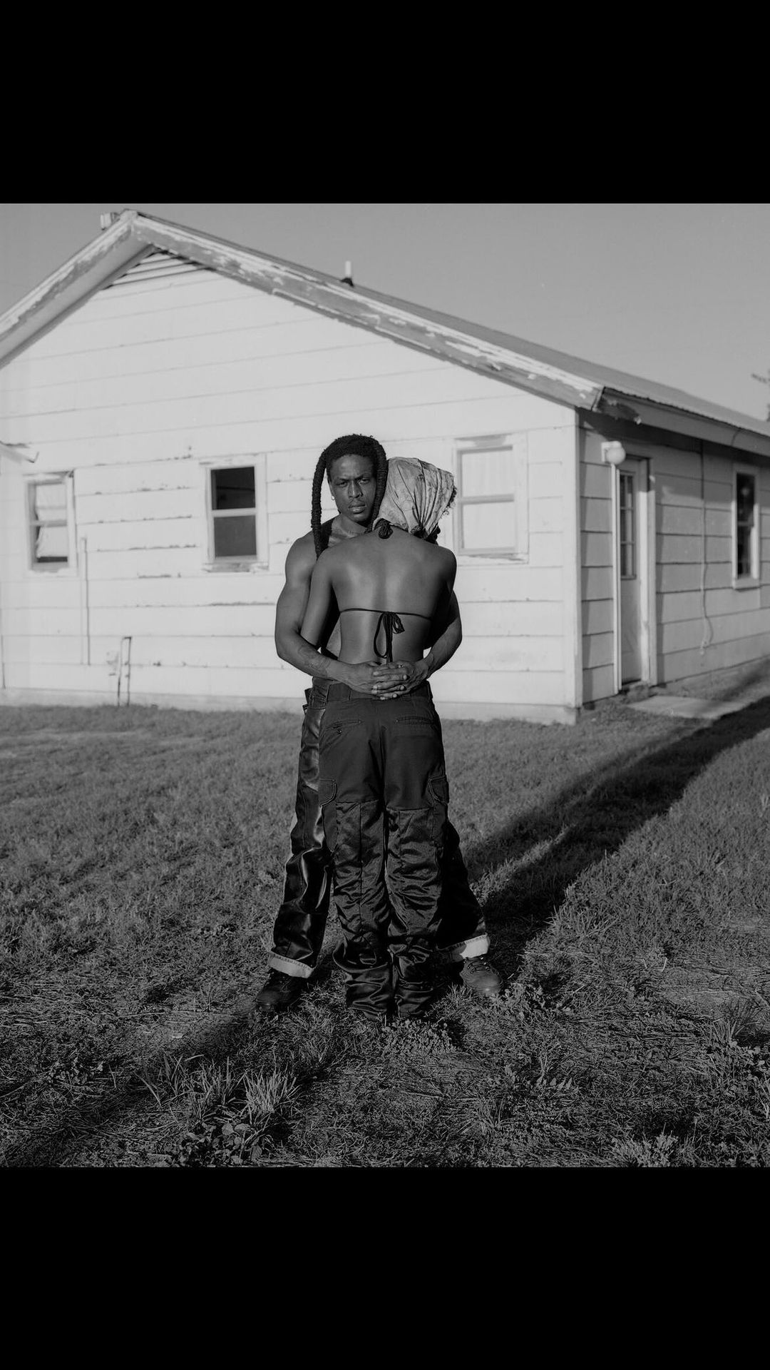 Black and white shot of two adults embracing in a field in front of a rural house on a sunny day, wearing loose clothing.