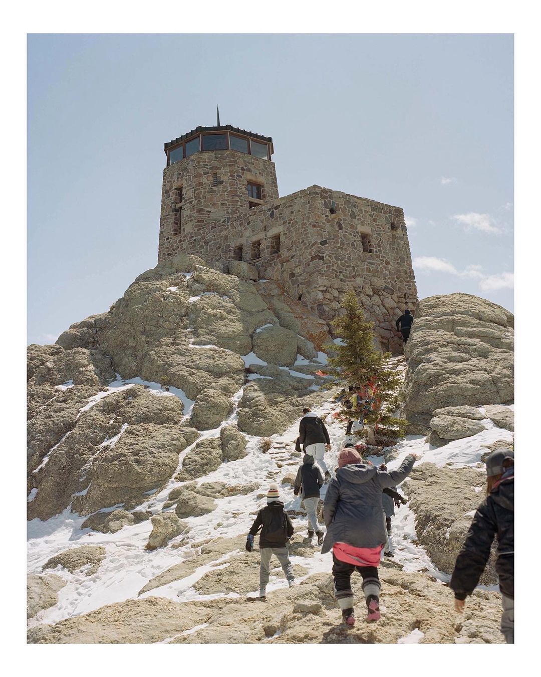 Stone fire lookout tower sits atop a mountain of boulders as a group of people walk up the rocky path towards the structure on a sunny day.