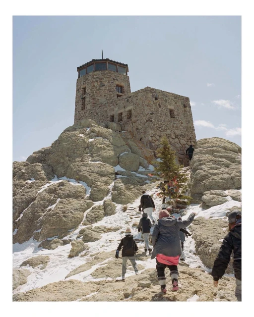 Stone fire lookout tower sits atop a mountain of boulders as a group of people walk up the rocky path towards the structure on a sunny day.