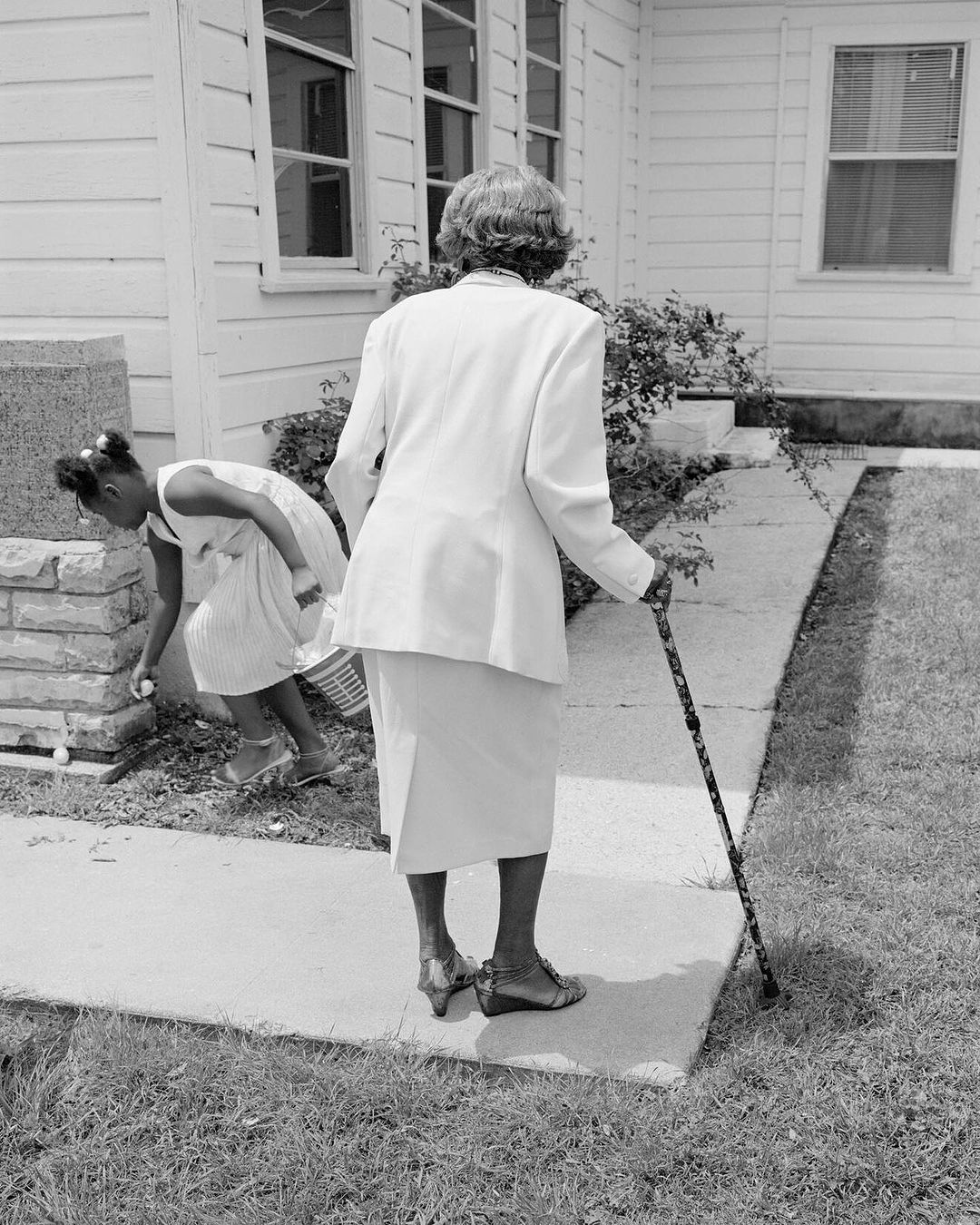Elderly woman in a white suit walks with a cane down a sidewalk, a girl in a dress gathers near a house, monochrome photograph.