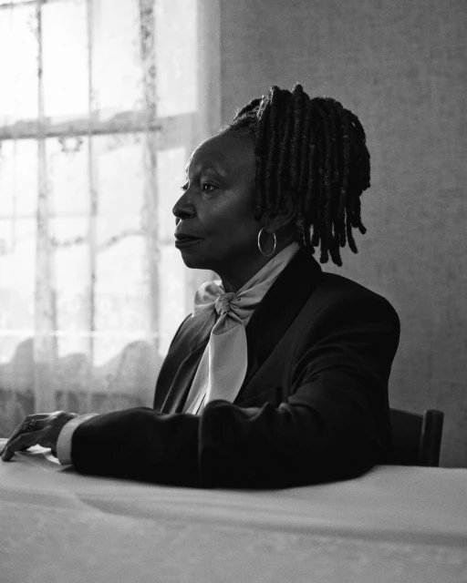 Black and white portrait of Whoopi Goldberg in suit, sitting at table by window with white lace curtains, looking left. Dreads hairstyle.