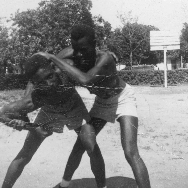 Two men wrestling outside, potentially in a park, captured in a black and white photograph with trees and a basketball hoop in the background.