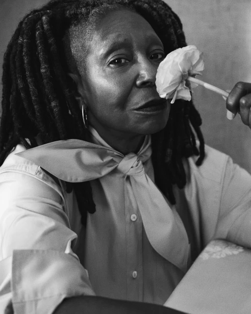 Black and white portrait of Whoopi Goldberg in collared shirt, posed with a white flower near her face, close-up shot.