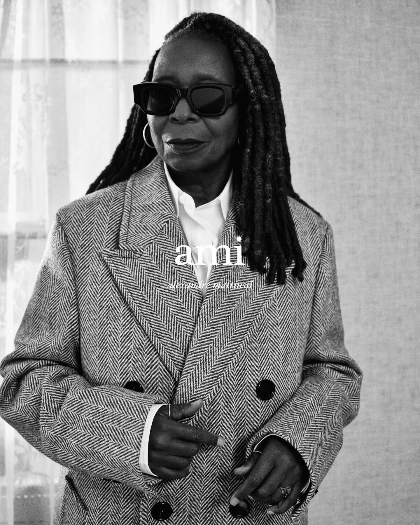 Black and white portrait of Whoopi Goldberg wearing dark sunglasses, a herringbone coat, and a white shirt, posing indoors.