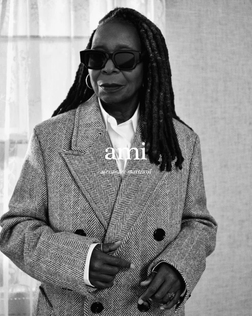 Black and white portrait of Whoopi Goldberg wearing dark sunglasses, a herringbone coat, and a white shirt, posing indoors.