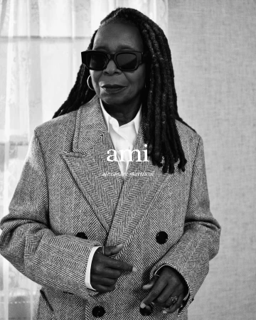 Black and white portrait of Whoopi Goldberg wearing dark sunglasses, a herringbone coat, and a white shirt, posing indoors.