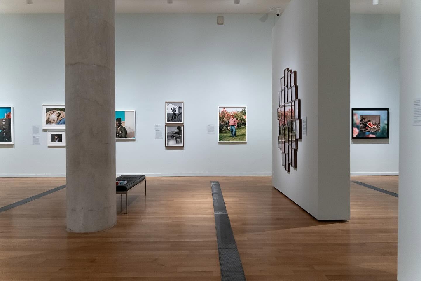 A gallery exhibition featuring framed photographs on white walls, with a polished wooden floor and a bench near a support column.