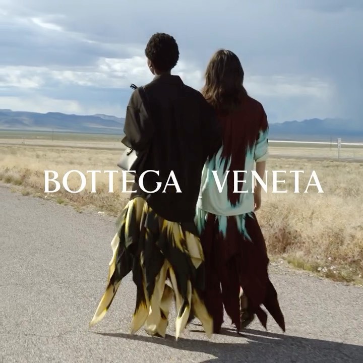 Two models in fashionable dresses walk on a remote road under a cloudy sky, showcasing Bottega Veneta's apparel and scenic landscapes.