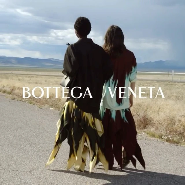 Two models in fashionable dresses walk on a remote road under a cloudy sky, showcasing Bottega Veneta's apparel and scenic landscapes.