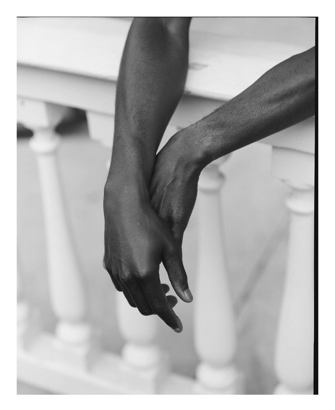 Black and white photo of a dark-skinned person's crossed arms resting on a white balustrade. Fingers are extended and downward.