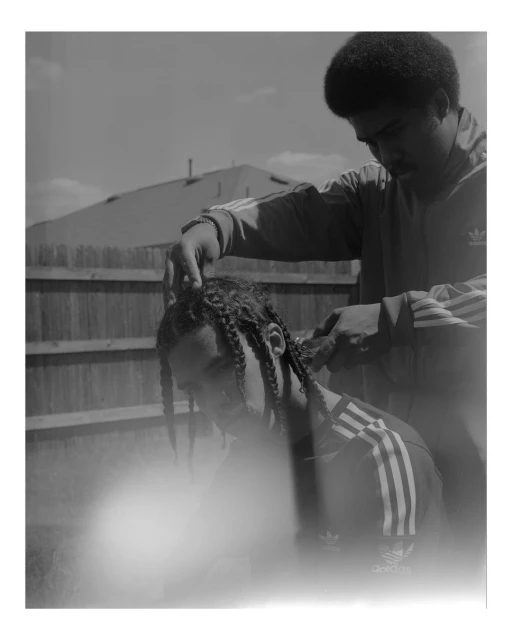 Two men outside, one braiding the other's hair. Both are wearing matching Adidas tracksuits with overcast sky and fence in background.