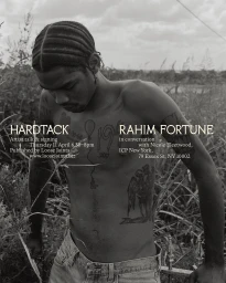 Black-and-white portrait of a shirtless young black man with tattoos outdoors among tall overgrown grasses.