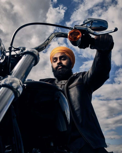 Man with beard and turban sits on a black motorcycle with a blue sky and white clouds in the background in a low-angle shot.