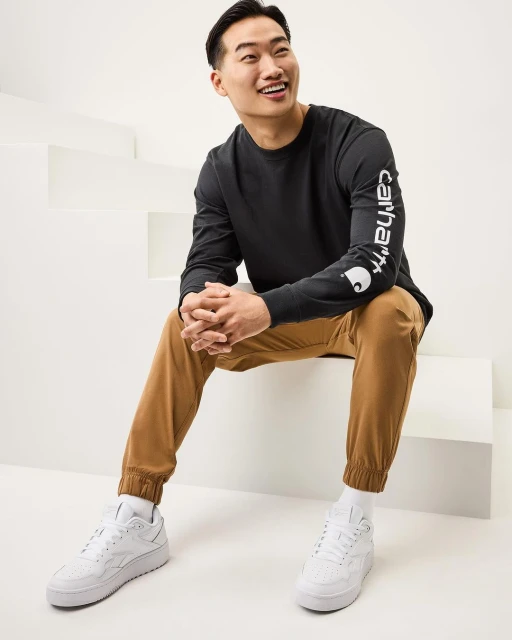 A smiling Asian man in a black Carhartt long-sleeve shirt and tan pants sits on white stairs wearing white sneakers in a studio shot.