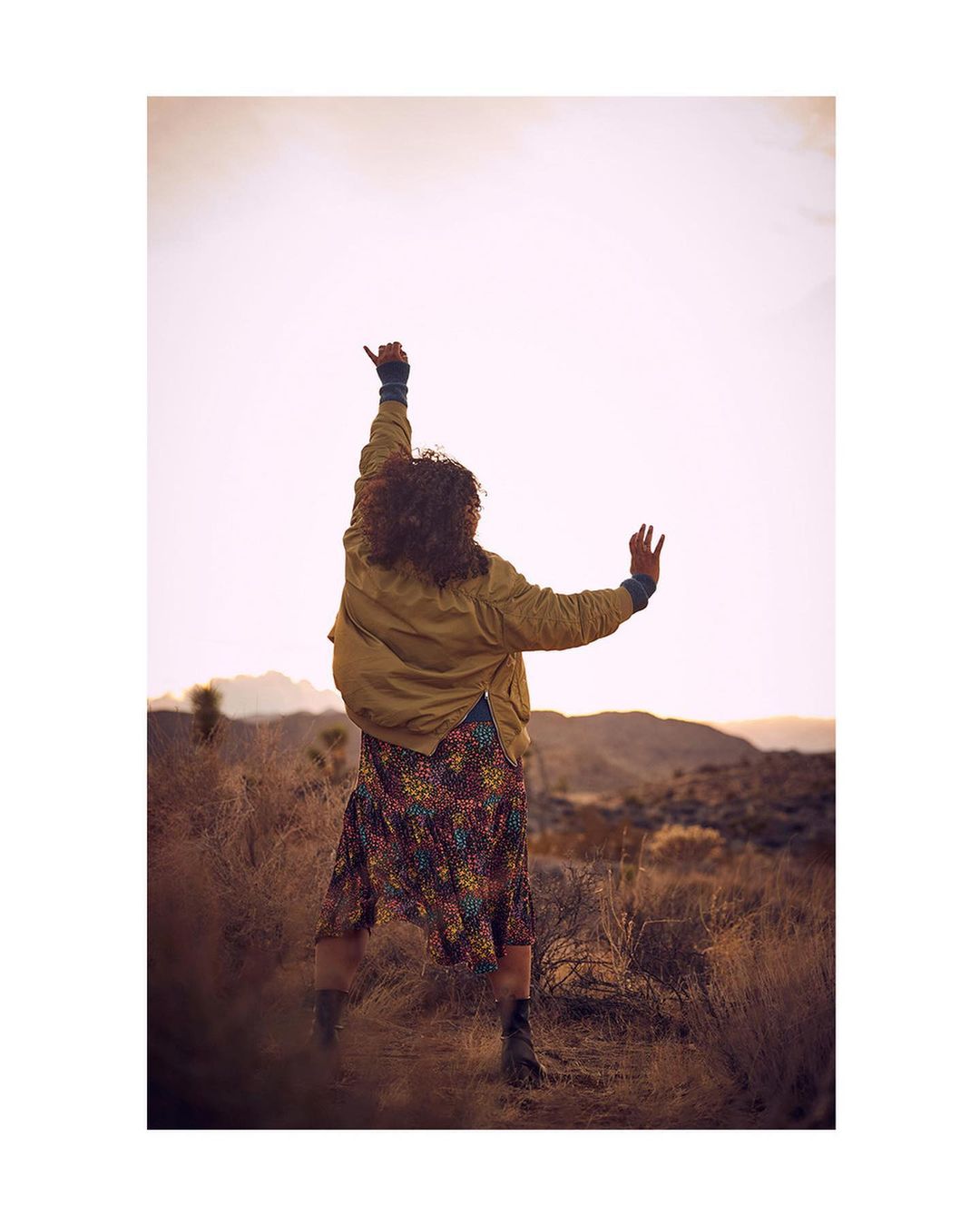 Woman in patterned dress and jacket stands in desert landscape with arms raised, back to camera, wearing boots. Outdoor, golden light.