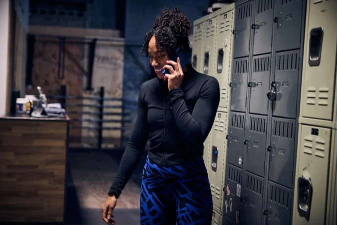 Woman smiles while talking on phone in gym locker room wearing blue patterned leggings, black top near gray lockers. Candid photo inside.