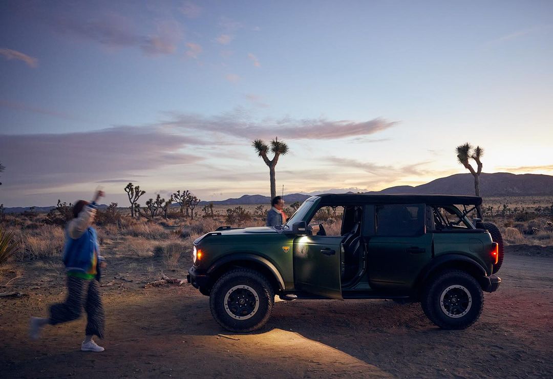 Off-road adventure with a green Ford Bronco in a desert landscape at sunset, with two people and sparse vegetation.