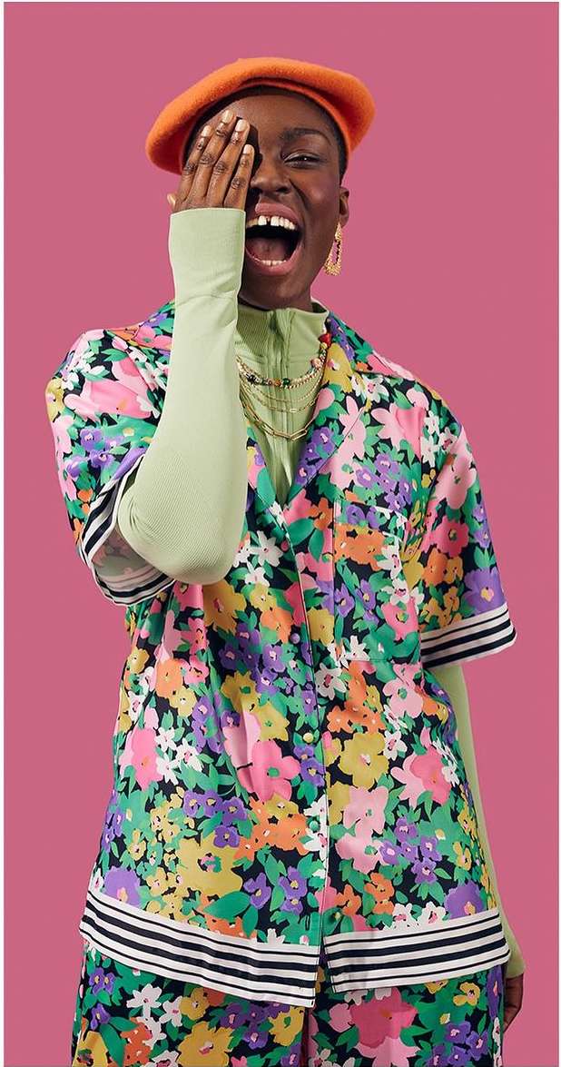Woman in floral shirt and orange beret covers her eye with her hand and smiles against a pink background in a medium studio shot.