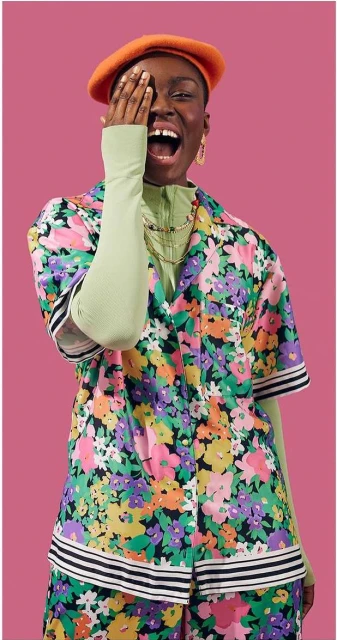 Woman in floral shirt and orange beret covers her eye with her hand and smiles against a pink background in a medium studio shot.