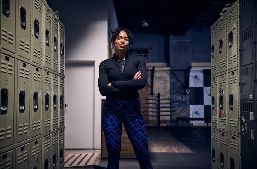A fit adult woman stands in a locker room with her arms crossed, wearing a black top and blue patterned leggings with a dark, moody atmosphere.
