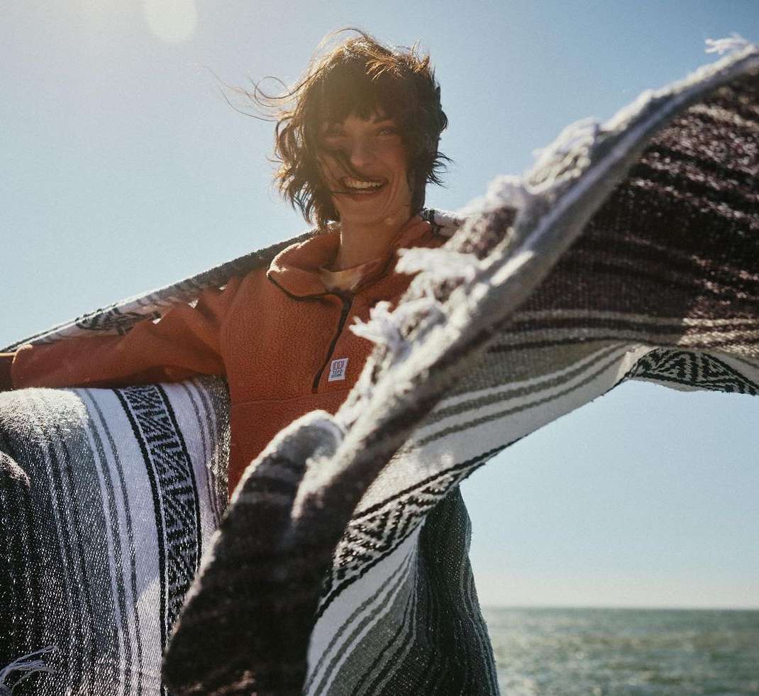 A smiling woman is wrapped in a colorful blanket outdoors on a bright day with a body of water visible in the background.