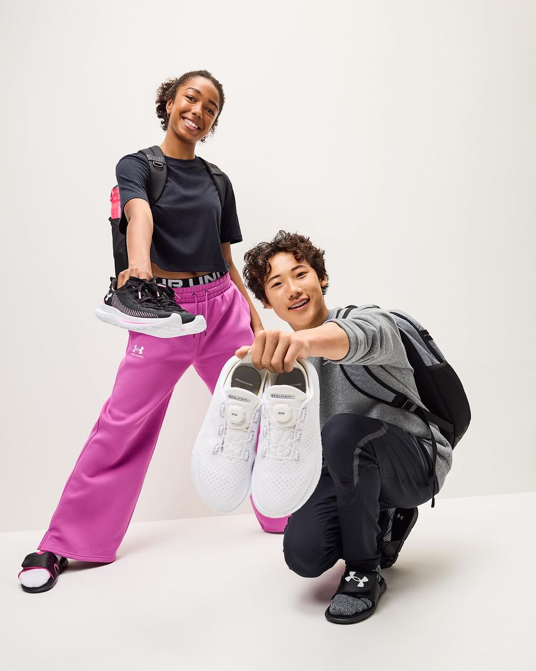 Two cheerful models hold Under Armour shoes against a clean white background, showcasing sporty athletic footwear and casual fashion.