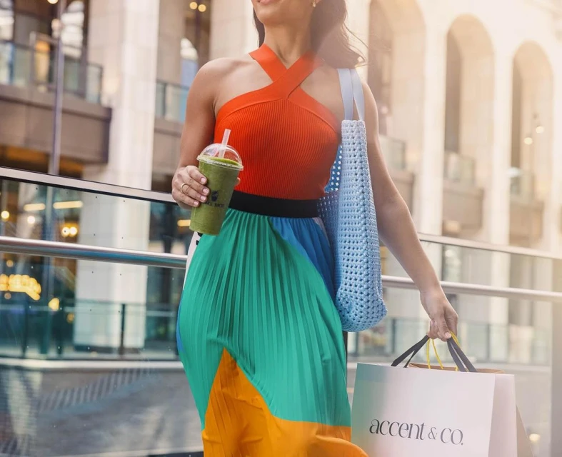 Woman walking in city with colorful skirt, holding green smoothie and shopping bags, buildings in background, medium outdoor shot.