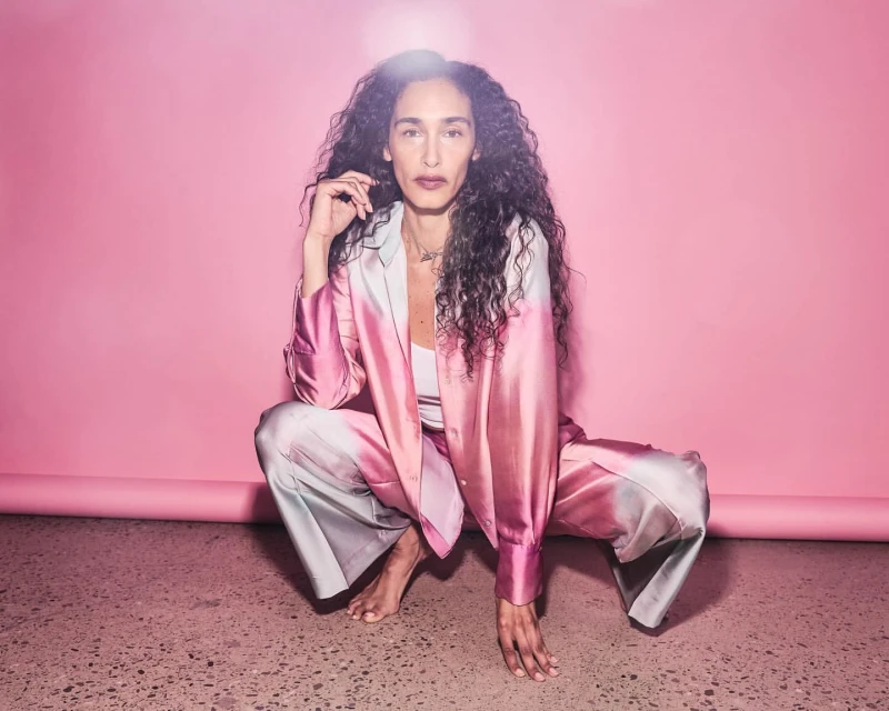 Attractive woman with long curly hair poses in a pink ombre suit against a pink backdrop. She is barefoot and crouching down.