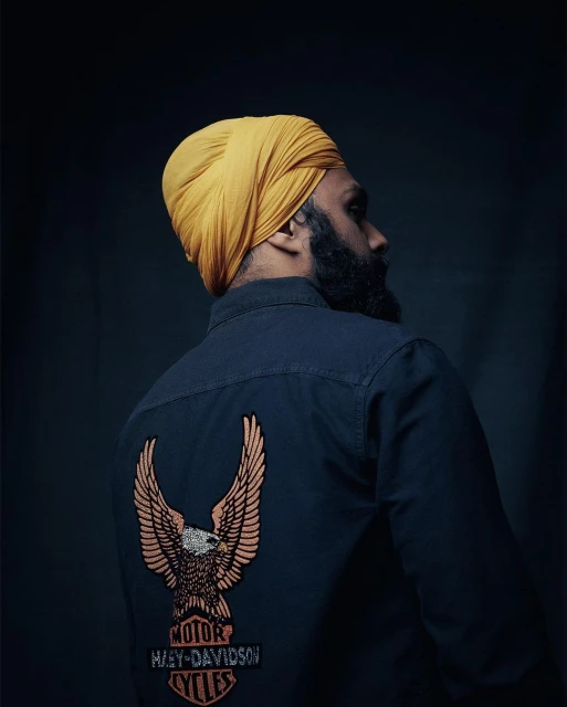 Portrait of a bearded adult male in a yellow turban, wearing a black shirt with a Harley-Davidson logo patch, against a dark background.