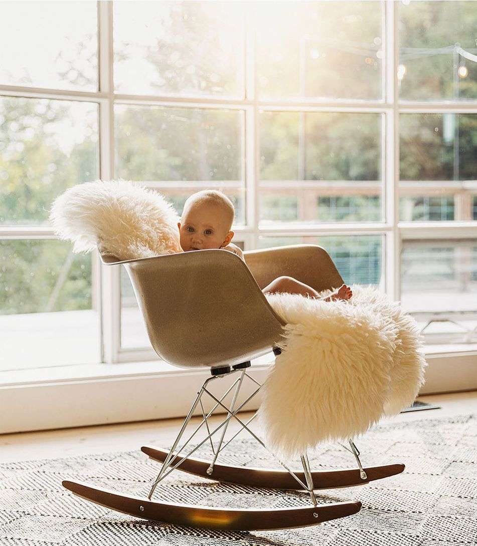 Infant sits in a rocking chair draped with a sheepskin rug in a bright room near a large window, creating a cosy, warm scene.