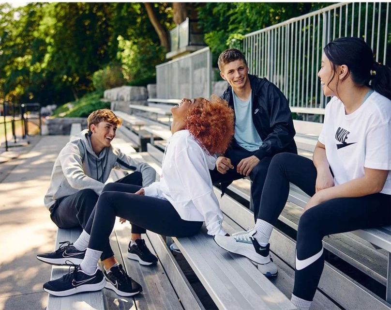 Four young adults wearing athletic apparel sit on stadium bleachers, socializing outdoors on a sunny day. They wear Nike branded clothing and shoes.