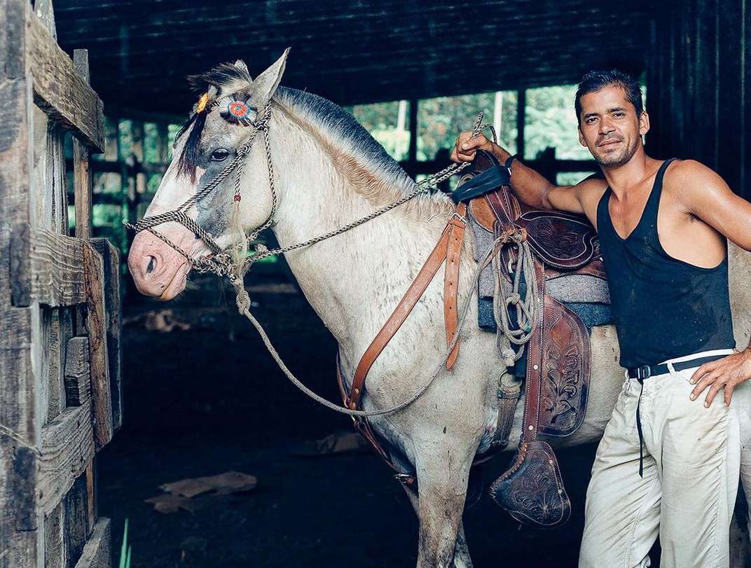 Tan adult male leans on a white horse with a saddle in a stable, with an open wooden door and green foliage visible in the background.